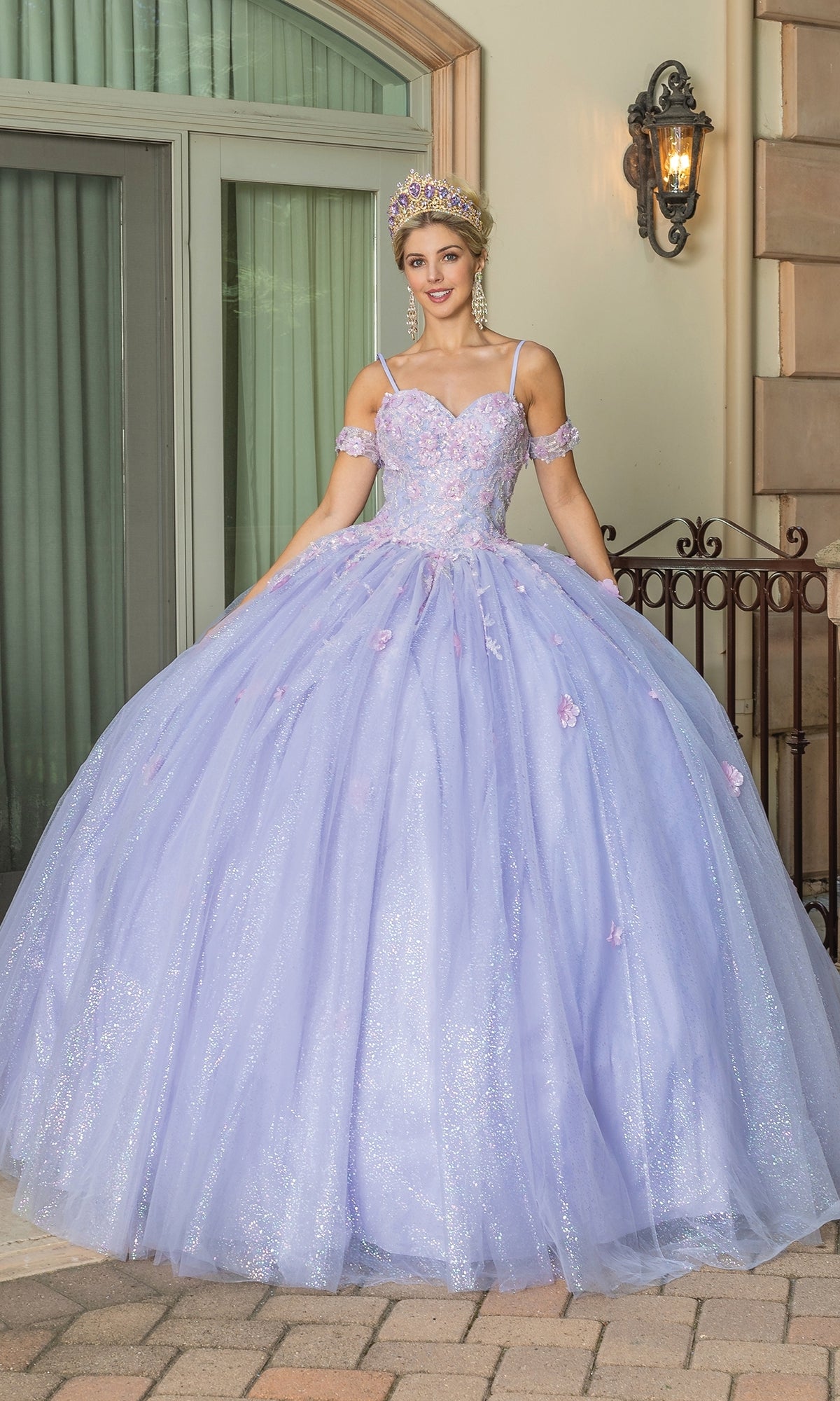 A young woman stands outdoors in front of a window, wearing the Quinceanera Dress 1785 By Dancing Queen—a sparkling lilac ball gown with a sweetheart neckline, floral details, plus a jeweled tiara and matching earrings.